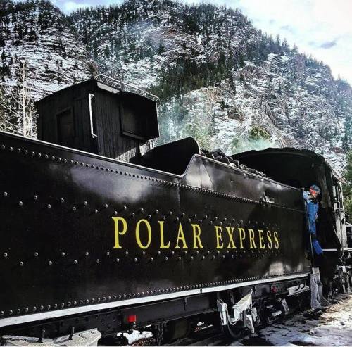 A black train labeled "Polar Express" near snowy mountains, with a conductor standing by the engine.