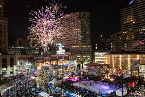 A vibrant city square filled with people celebrating, fireworks lighting up the night sky above an ice skating rink.