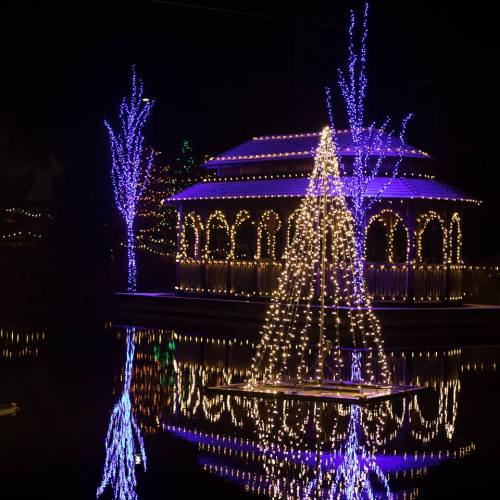 A beautifully lit gazebo surrounded by colorful lights, reflecting on a calm water surface at night.