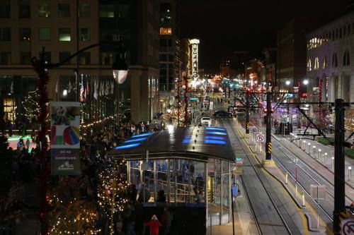A bustling city street at night, decorated for the holidays, with a tram station and festive lights in the background.