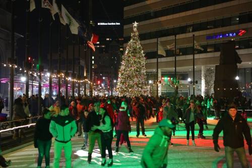 A bustling ice skating rink with a large Christmas tree, surrounded by festive lights and a crowd of skaters.