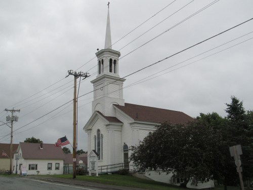 A white church with a tall steeple, surrounded by trees, under a cloudy sky, with an American flag nearby.