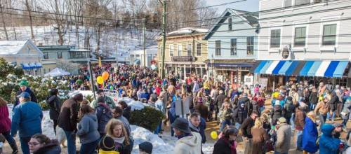 A large crowd gathers in a snowy town square, surrounded by shops and festive decorations on a sunny day.