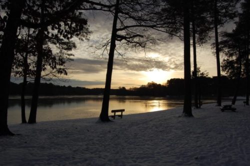A serene winter landscape with a snowy shore, trees, and a calm lake reflecting a sunset sky.