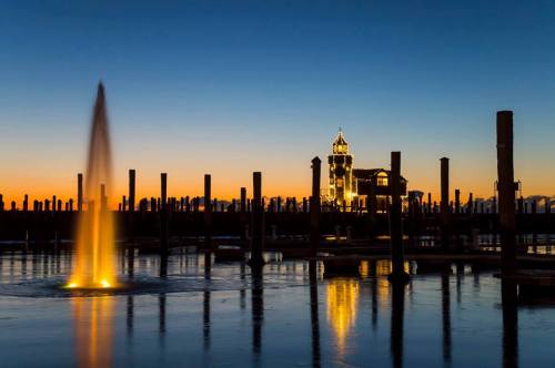 A serene sunset over a calm waterway, featuring a fountain and silhouetted pilings with a lit building in the background.