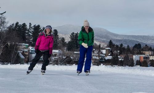 Two women ice skating on a frozen lake, surrounded by trees and mountains, wearing colorful winter jackets.