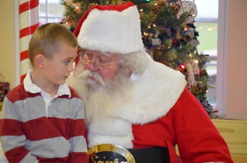 A young boy in a striped shirt sits closely with Santa Claus, who has a white beard and a red suit, in a festive setting.