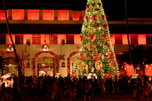 A festive scene with a large decorated Christmas tree and crowds of people enjoying holiday decorations at night.