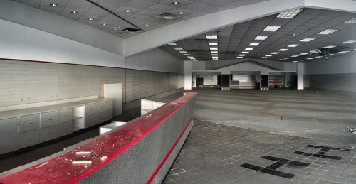 An empty, abandoned retail space with gray walls, a red counter, and scattered debris on the floor.
