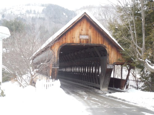 A snow-covered wooden covered bridge surrounded by trees and mountains in winter.
