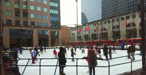 A busy outdoor ice skating rink surrounded by buildings, with children and adults skating and flags in the background.