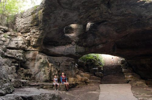A woman and two children sit on rocks under a large stone archway, with stairs leading up in the background.