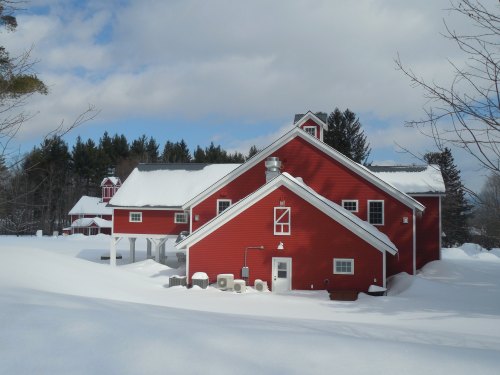 A red barn surrounded by snow, with a cloudy sky and trees in the background. Winter scene in a rural setting.