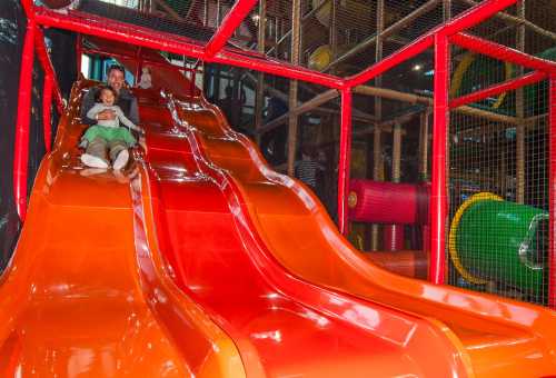 A child and an adult slide down bright orange slides in a colorful indoor play area.