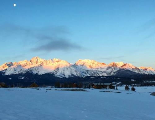 Snow-covered mountains bathed in golden light at sunset, with a clear blue sky and a hint of the moon.