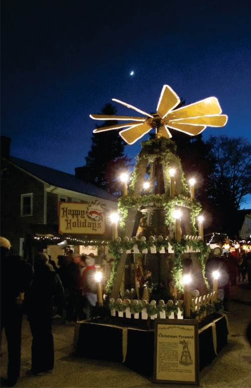 A festive Christmas pyramid adorned with lights and greenery, surrounded by people at a holiday market under a night sky.