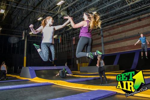 Two women jump on trampolines at a trampoline park, with others bouncing in the background. Bright and energetic atmosphere.
