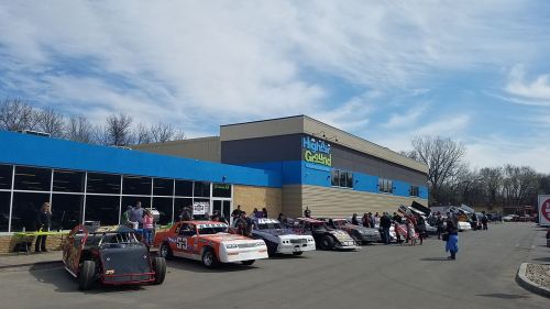 A line of race cars parked outside a blue and gray building on a sunny day, with people gathered nearby.
