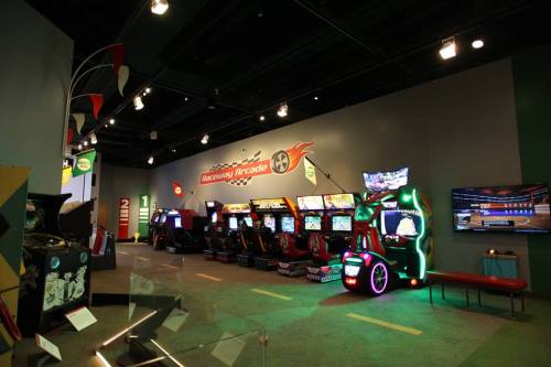 A vibrant arcade with various gaming machines and a large screen, featuring racing-themed decor and bright lighting.