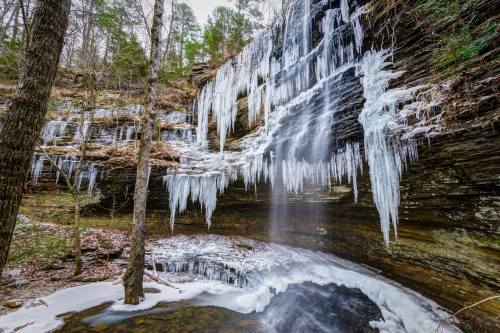 A frozen waterfall with icicles hanging from rocky cliffs, surrounded by trees and a snowy landscape.