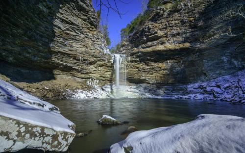A serene waterfall cascades into a snowy, rocky pool surrounded by steep, icy cliffs under a clear blue sky.