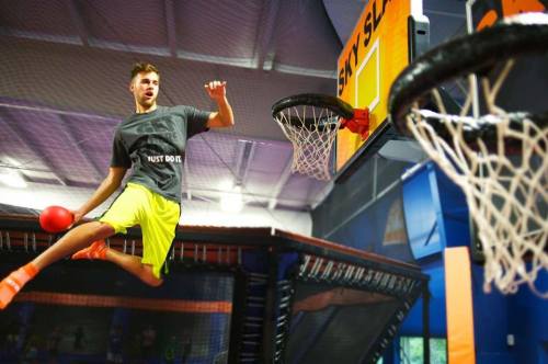 A young man jumps in mid-air, preparing to dunk a basketball at a trampoline park. Bright colors and energetic atmosphere.
