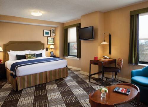 Cozy hotel room featuring a bed, desk, chair, and TV, with warm colors and natural light from windows.