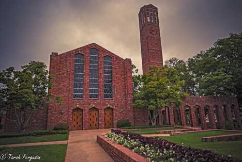 A brick building with large windows and a clock tower, surrounded by trees and flower beds under a cloudy sky.