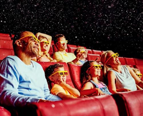 A group of people, including children, enjoy a movie in a theater, wearing 3D glasses and smiling as snow falls.