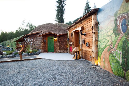 A whimsical wooden house with a thatched roof, green door, and colorful mural, surrounded by trees and gravel path.