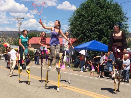 A group of performers on stilts entertain a crowd at a street festival under a blue sky with fluffy clouds.
