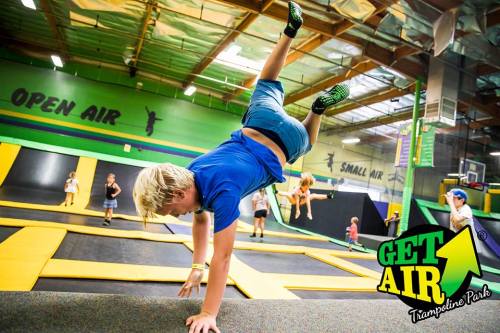 A child performs a handstand on a trampoline at a vibrant indoor trampoline park, with others jumping in the background.