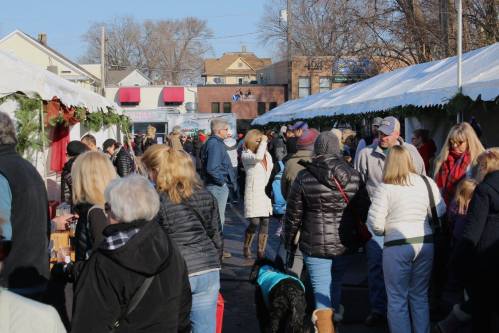A bustling outdoor market scene with many people browsing stalls under white tents on a sunny day.