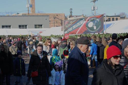 A busy outdoor market scene with many people walking among tents and stalls, surrounded by greenery and buildings.
