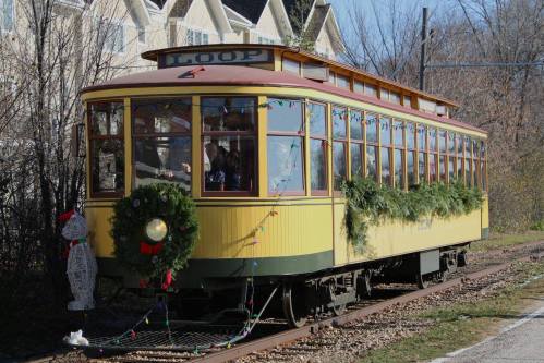 A vintage yellow streetcar decorated with holiday wreaths and lights, parked along a tree-lined track.