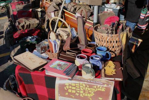A display of handmade crafts, including mugs, notebooks, and woven baskets, on a checkered tablecloth at a market.