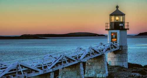 A lighthouse stands on a rocky shore at sunset, with calm waters and distant hills in the background.