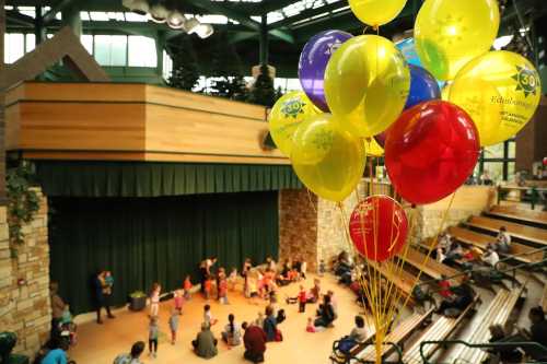 Colorful balloons in the foreground with children performing on stage in a spacious indoor venue.