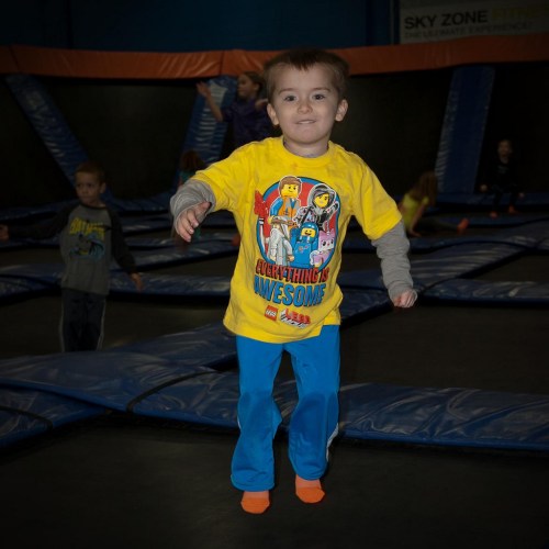 A young boy in a bright yellow shirt jumps on a trampoline, smiling, with other children playing in the background.