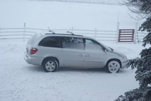 A silver minivan parked in deep snow, with a snowy landscape and a white fence in the background.
