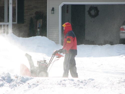 A person in a red and blue jacket uses a snowblower to clear snow from a driveway on a winter day.