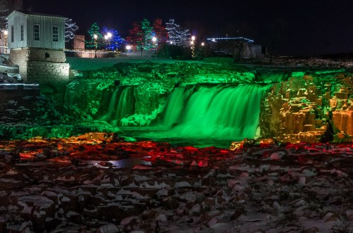 A waterfall illuminated in green and red lights, surrounded by snow and festive decorations at night.