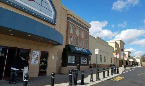 A shopping plaza with various storefronts and a clear blue sky, featuring a movie theater and restaurants.
