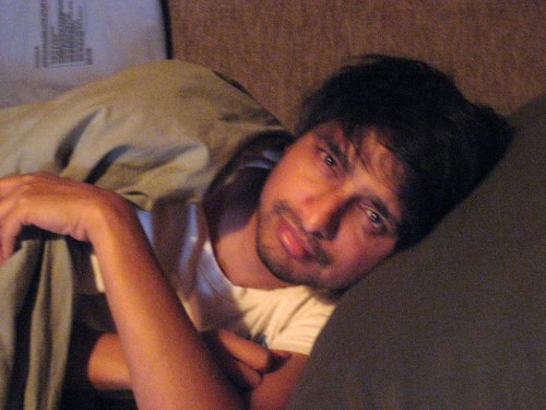 A person lying in bed, partially covered with a blanket, looking directly at the camera with a relaxed expression.