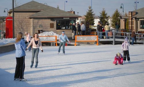 A group of people ice skating on a rink, with spectators in the background enjoying the winter day.