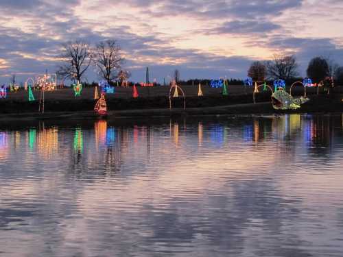 Colorful holiday lights reflect on a calm river at sunset, with trees and decorations lining the shore.