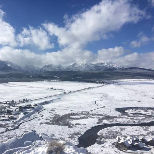 A snowy landscape with mountains in the background, a winding river, and scattered houses under a partly cloudy sky.