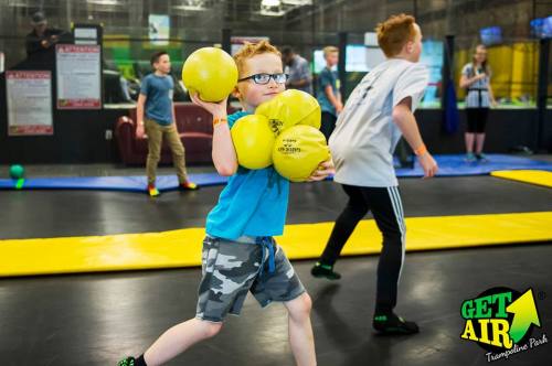A young boy holds two yellow balls, preparing to throw, in a trampoline park with other kids playing in the background.