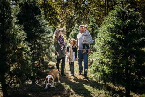 A family with two children and a dog stands among evergreen trees, enjoying a sunny day outdoors.