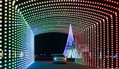 A car drives through a colorful light tunnel, with a decorated Christmas tree in the background.